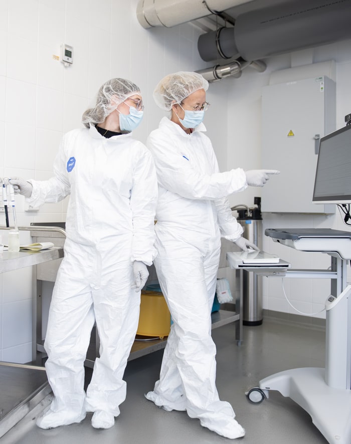 Michèle Lindner and a laboratory technician in protective suits and masks discussing in front of a screen in the cleanroom.